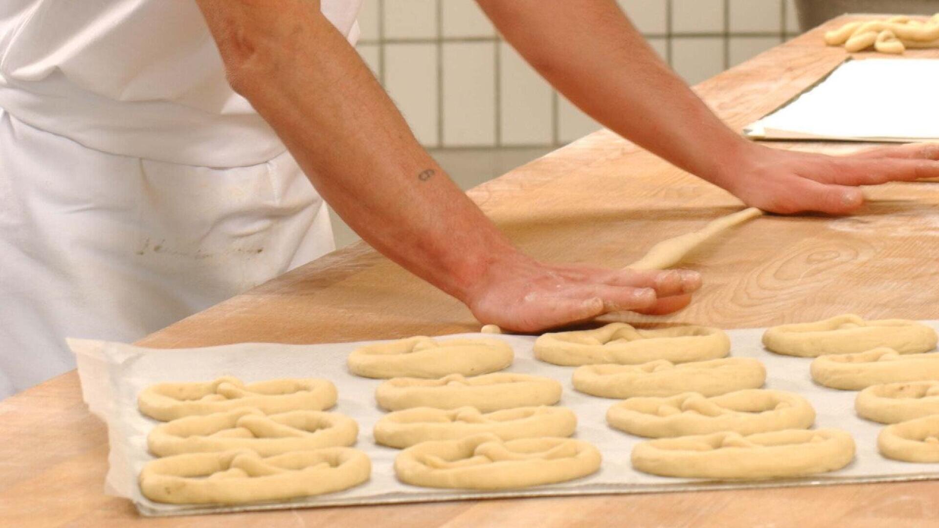 A person in a white apron shapes dough on a wooden table, preparing pretzels. Several unbaked pretzels are arranged on parchment paper, ready for baking.
