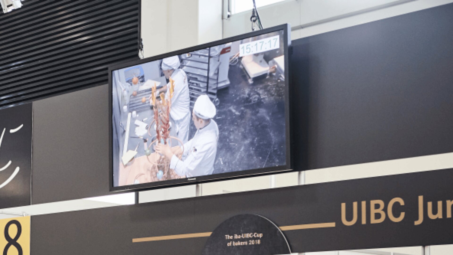 Ein Monitor zeigt eine Live-Videoübertragung von drei Konditoren in weißen Uniformen, die in einer Küche an einer großen, komplizierten Schokoladenskulptur arbeiten. Die Uhrzeit auf dem Bildschirm lautet 15:17:17.