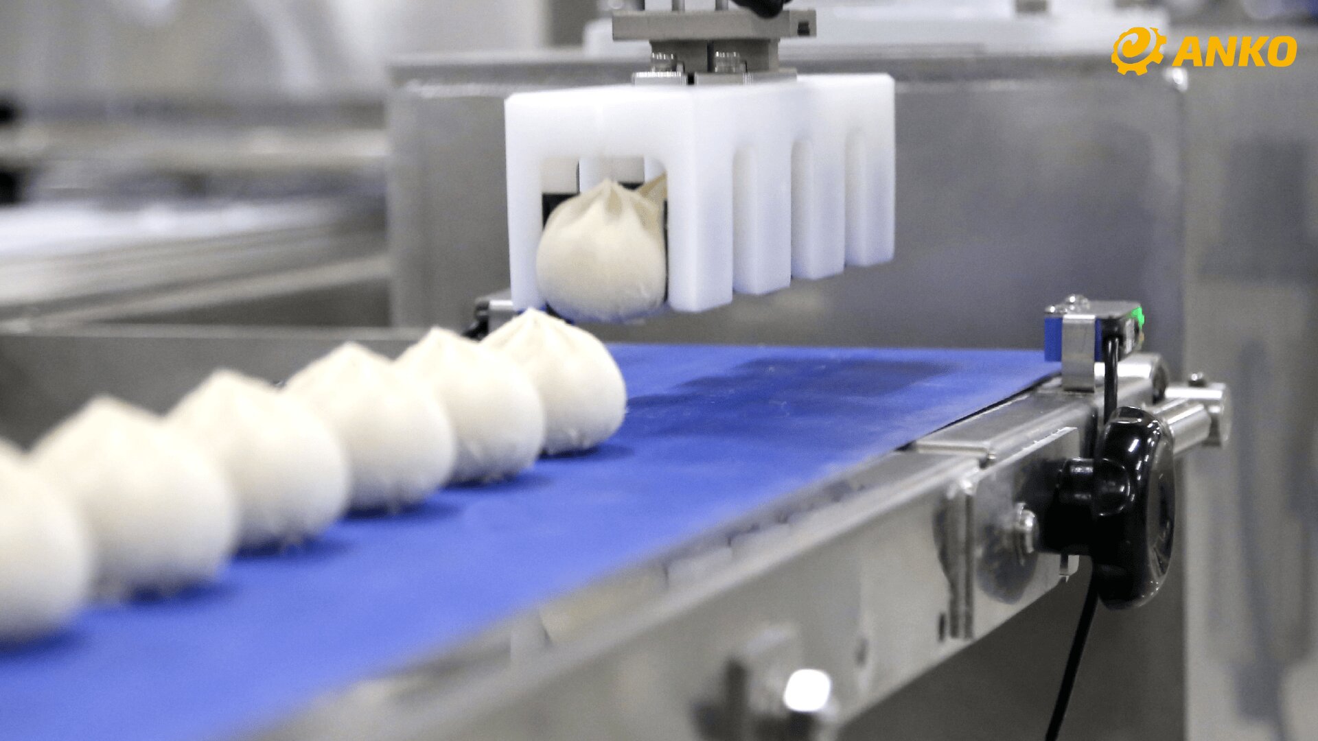 A row of steamed buns moves along a blue conveyor belt in a food processing factory, with a white mechanical device handling one bun. The ANKO logo is visible in the top right corner.