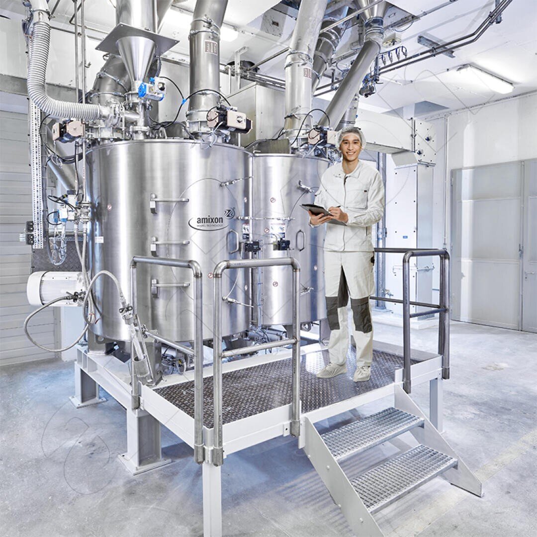 A worker in a white uniform and cap stands on a platform next to large stainless steel industrial mixing tanks in a clean, bright factory setting, holding a tablet and smiling at the camera.