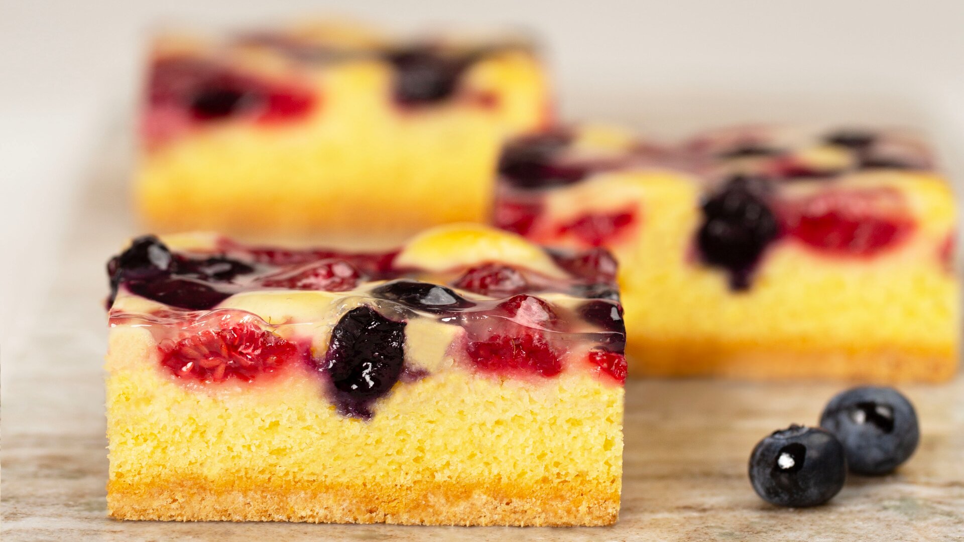 Nahaufnahme eines quadratischen Stücks gelben Biskuitteigs, das mit einer klaren Glasur, Himbeeren und Blaubeeren belegt ist. Zwei frische Blaubeeren sind in der Nähe auf der Oberfläche platziert. Weitere Kuchenscheiben sind im Hintergrund unscharf.
