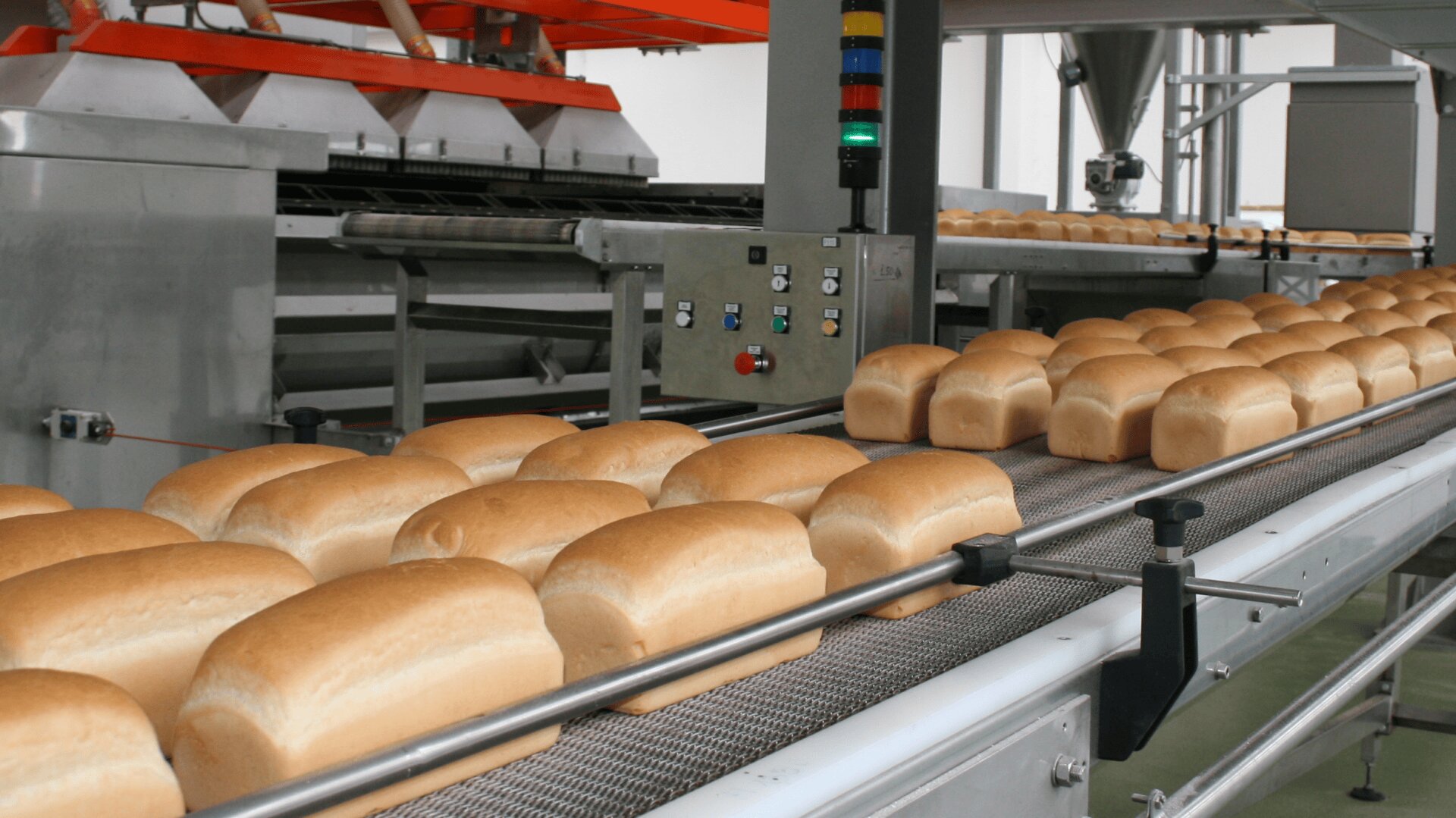 Loaves of bread move along a conveyor belt in an industrial bakery, with machinery and control panels visible in the background.