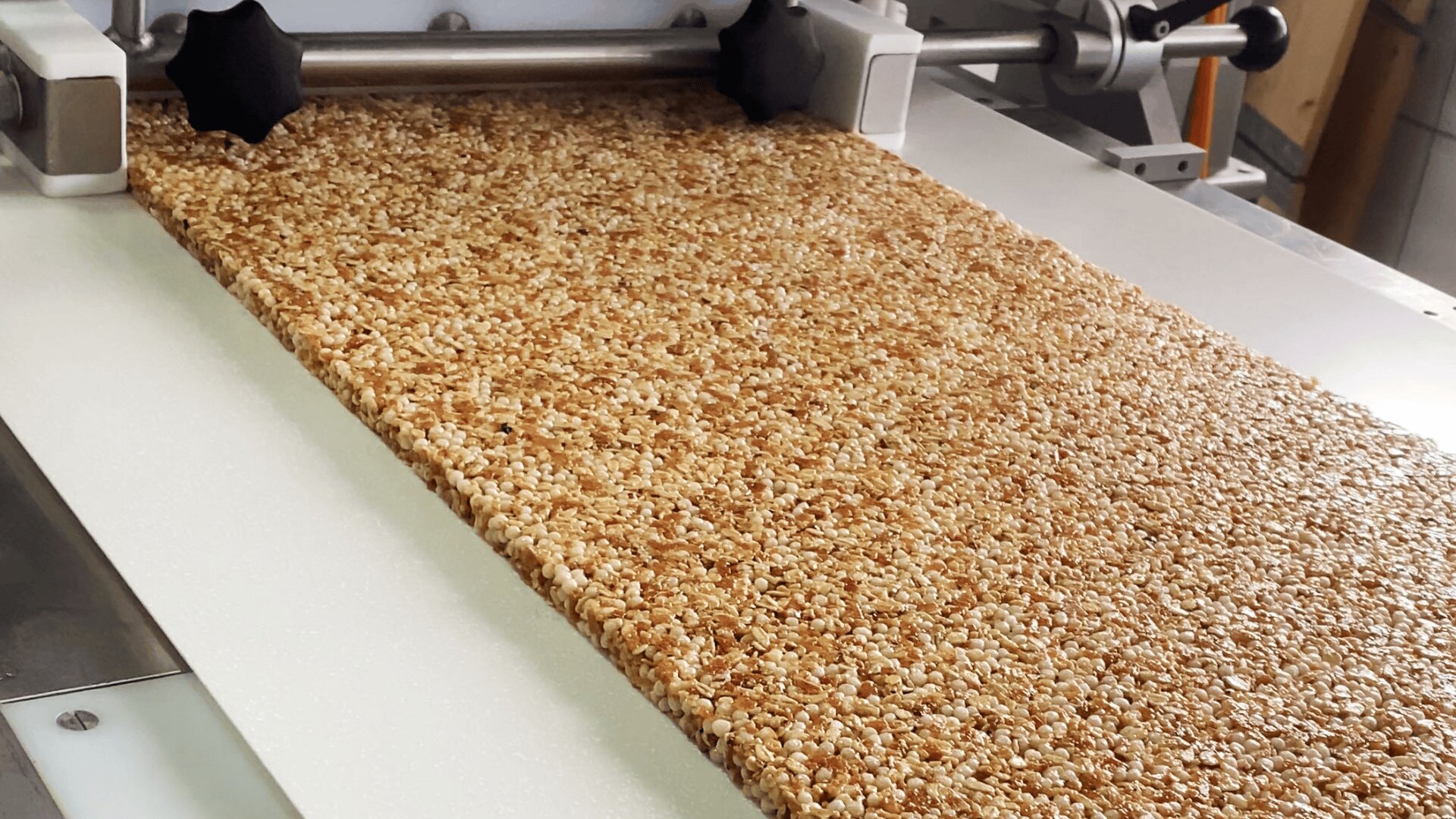 A large sheet of granola is being processed by an industrial machine, likely being flattened and shaped before cutting into bars. The mixture appears golden brown with visible oats and seeds.