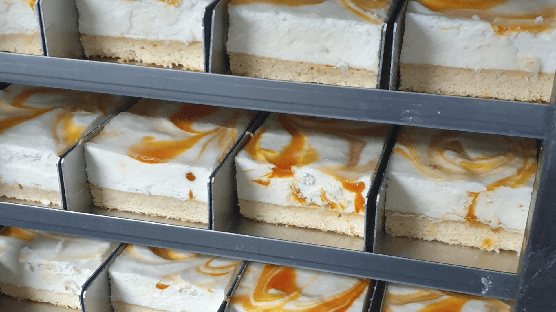 In Scheiben geschnittene Dessertriegel mit einem krümeligen Boden, einer cremigen weißen Schicht und einem orangefarbenen Karamellstrudel obenauf, ordentlich in Metallschalen angeordnet.