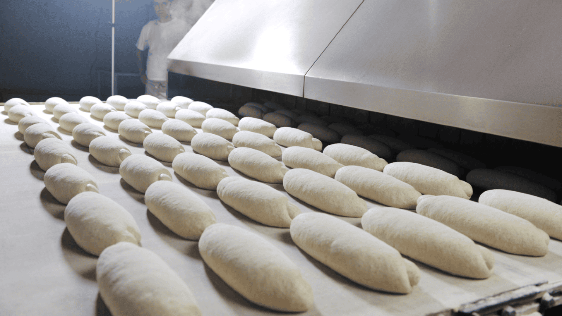 Rows of unbaked bread loaves on a conveyor belt moving into an industrial oven, with a worker standing in the background amidst steam.
