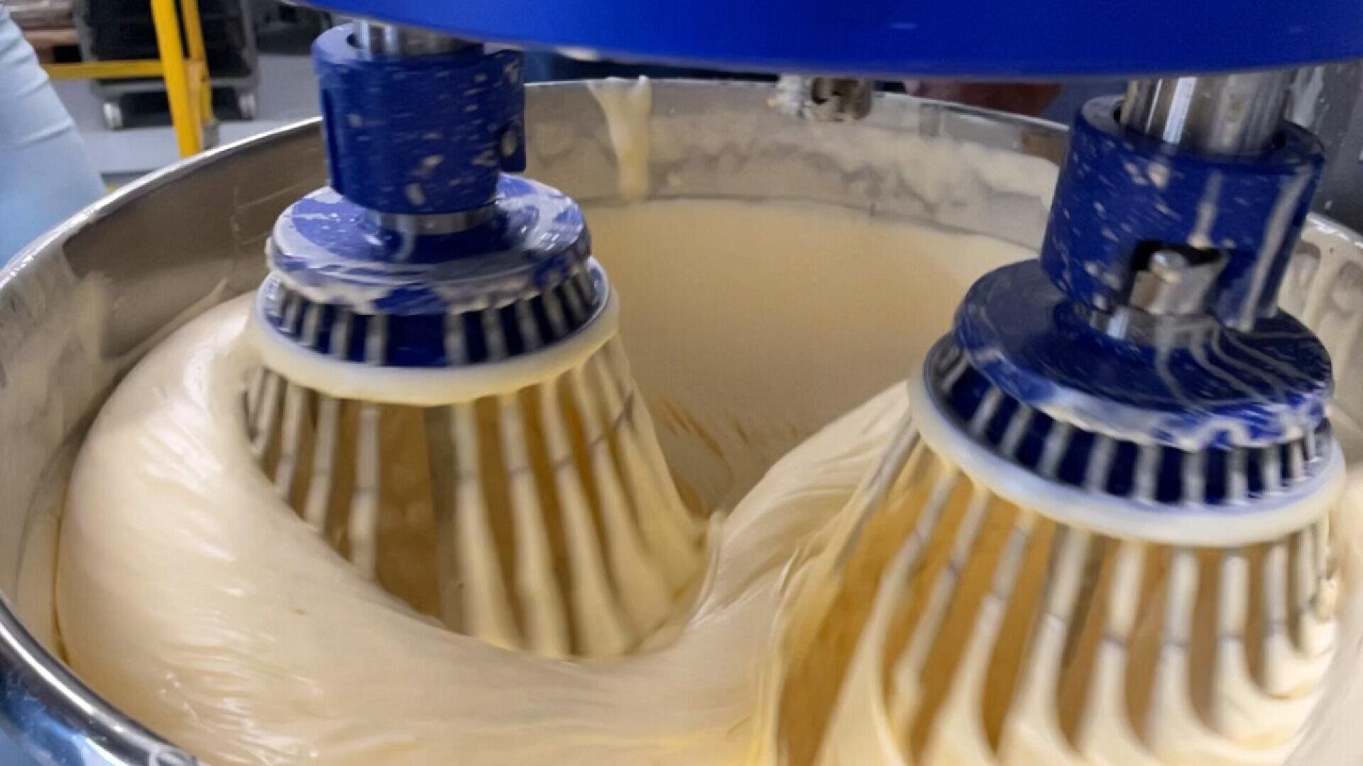 Nahaufnahme eines industriellen Mixers mit zwei großen blauen Quirlen, die einen dicken, cremigen Teig in einer Metallschüssel verrühren. Der Teig haftet an den sich drehenden Rührbesen, während sie sich drehen.