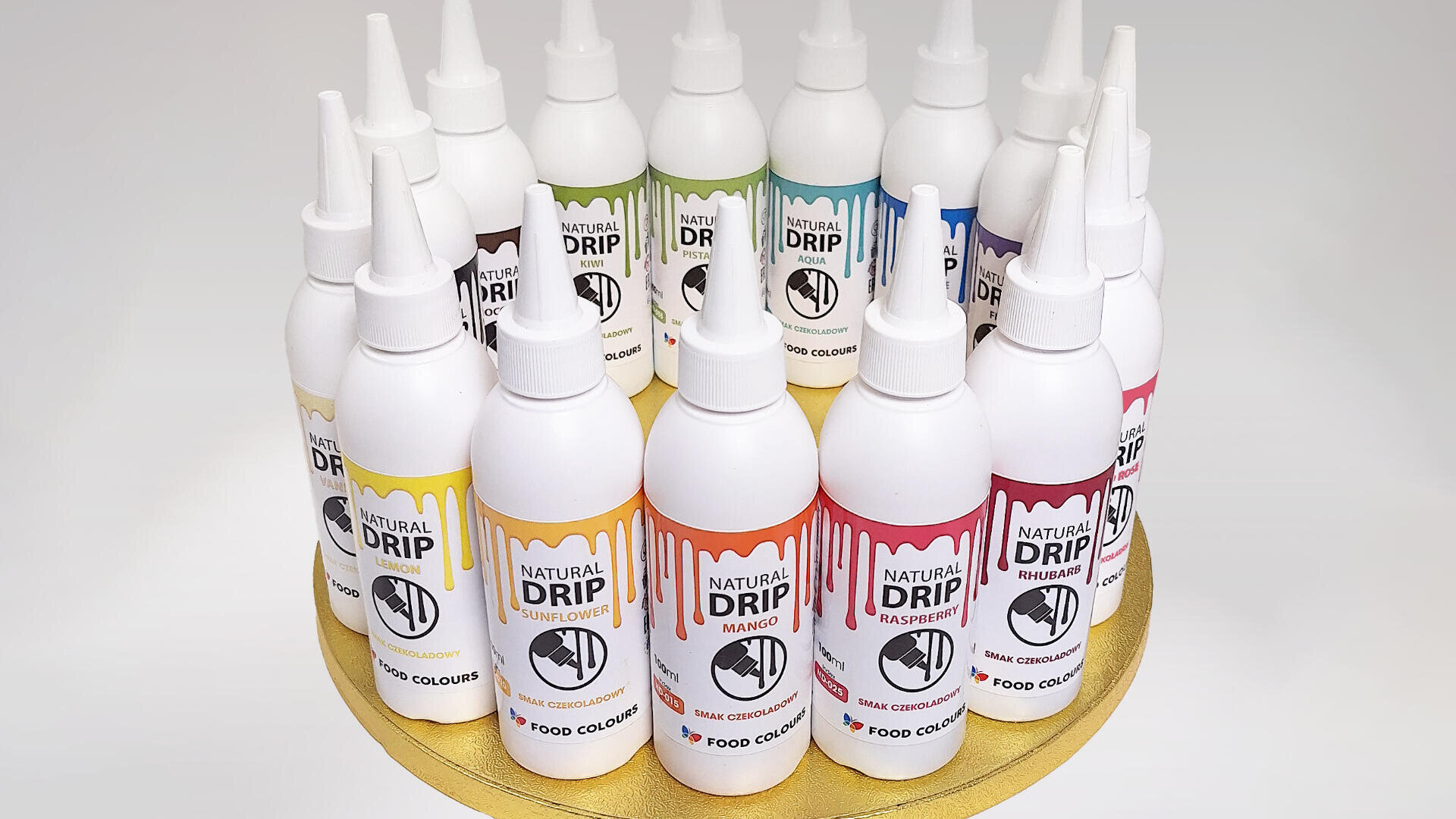 Several bottles of Natural Drip food colouring are arranged in a circle on a gold surface. Each bottle has a different colour and flavor label, such as mango, raspberry, and passion fruit.