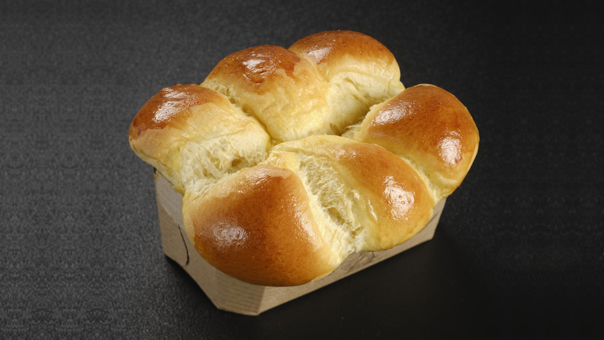 A golden, glossy loaf of pull-apart bread with six soft, fluffy rolls sits in a small brown paper tray on a dark surface.