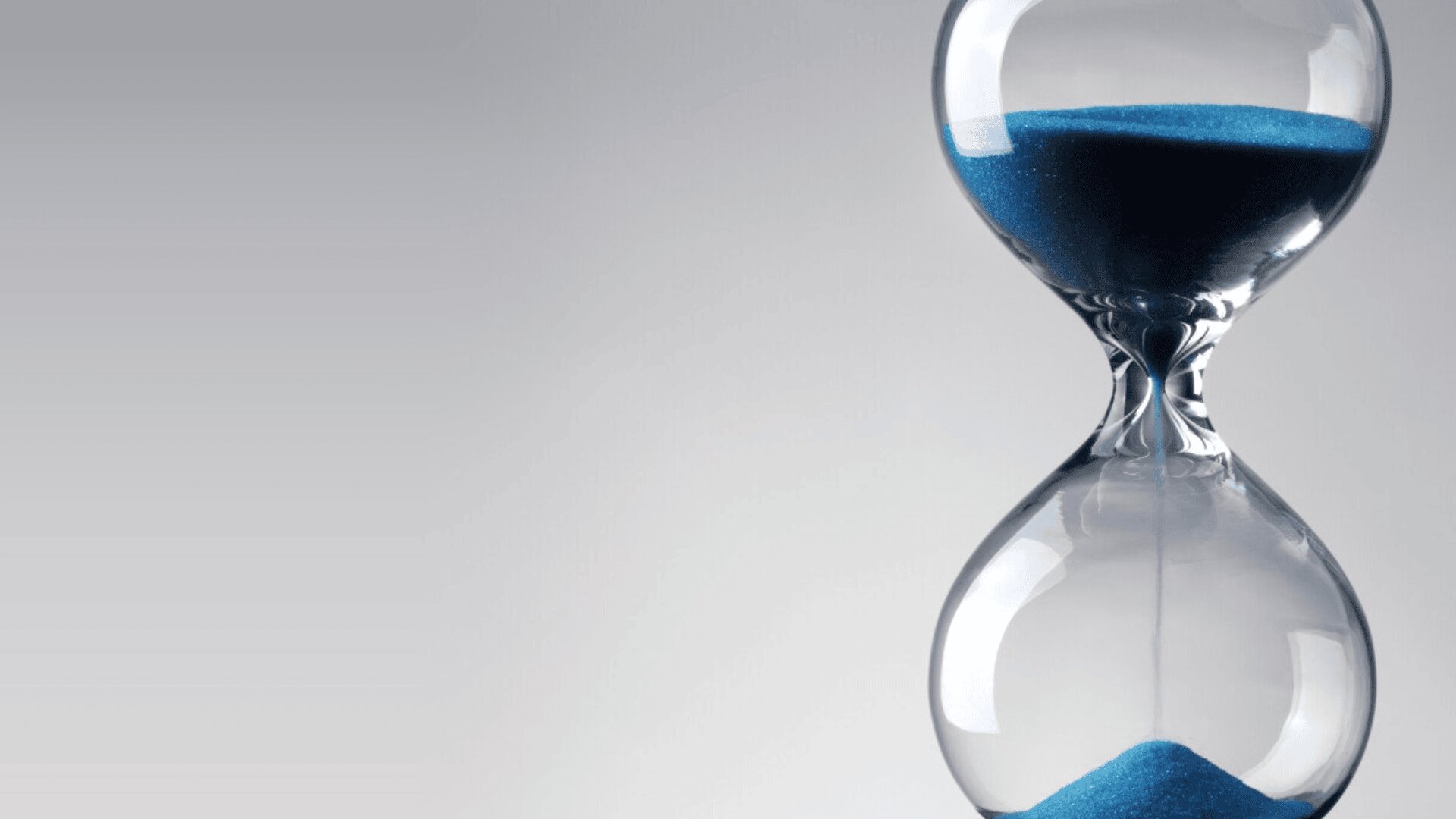 A close-up of a glass hourglass with blue sand falling from the upper chamber to the lower chamber, set against a light gray background.