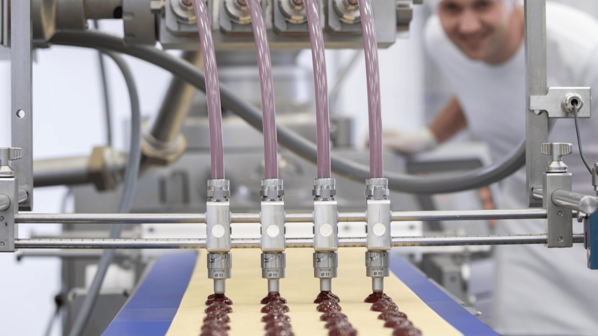 Eine Lebensmittelverarbeitungsmaschine gibt Reihen von Gelee oder Marmelade auf ein Förderband ab, wobei ein lächelnder Arbeiter in weißer Uniform unscharf im Hintergrund zu sehen ist.
