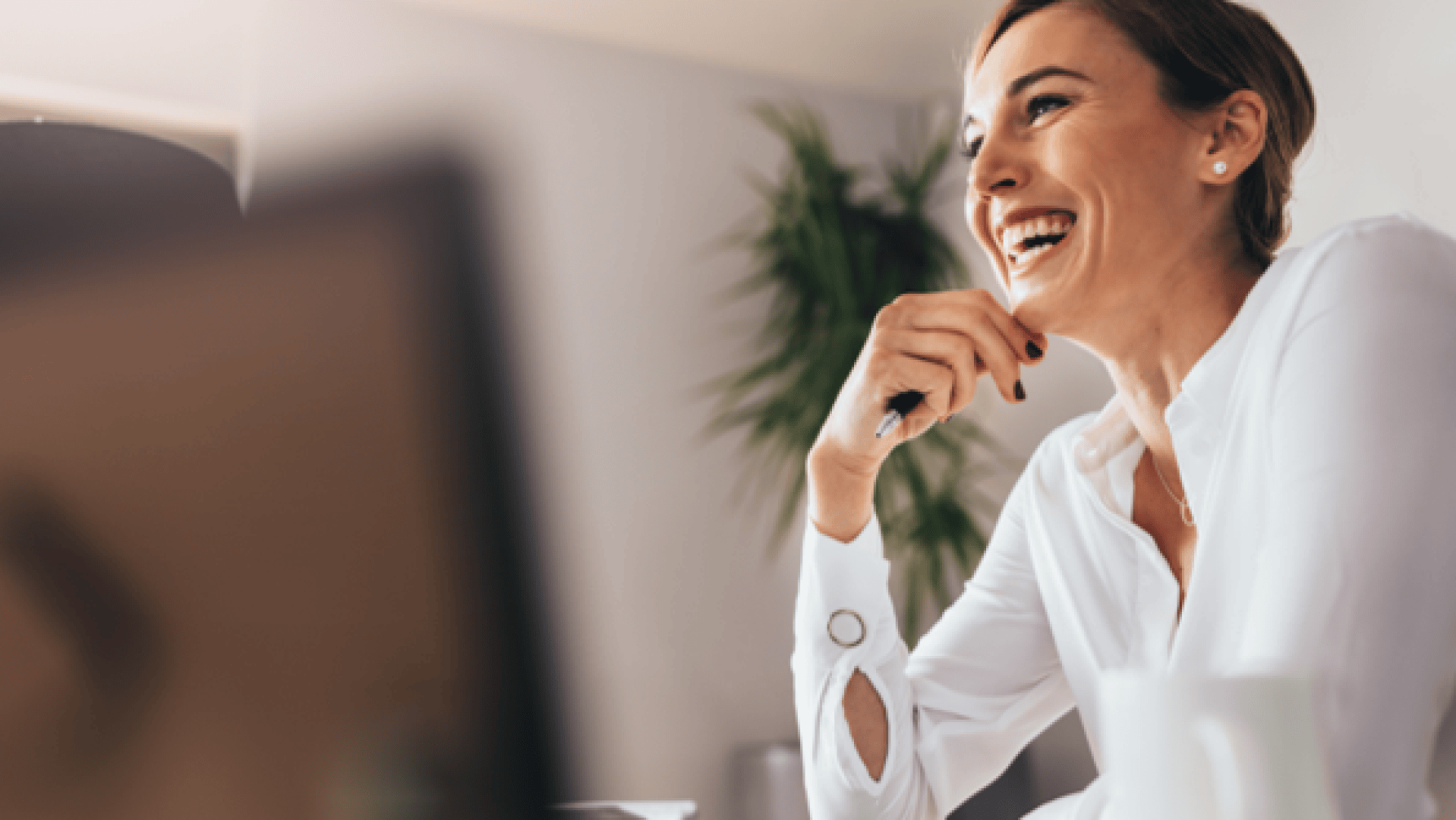 Eine lächelnde Frau in weißer Bluse sitzt an einem Schreibtisch, hält einen Stift in der Hand und blickt zur Seite, im Vordergrund sind eine Kaffeetasse und ein unscharfer Computerbildschirm zu sehen. Im Hintergrund ist eine Grünpflanze zu sehen.