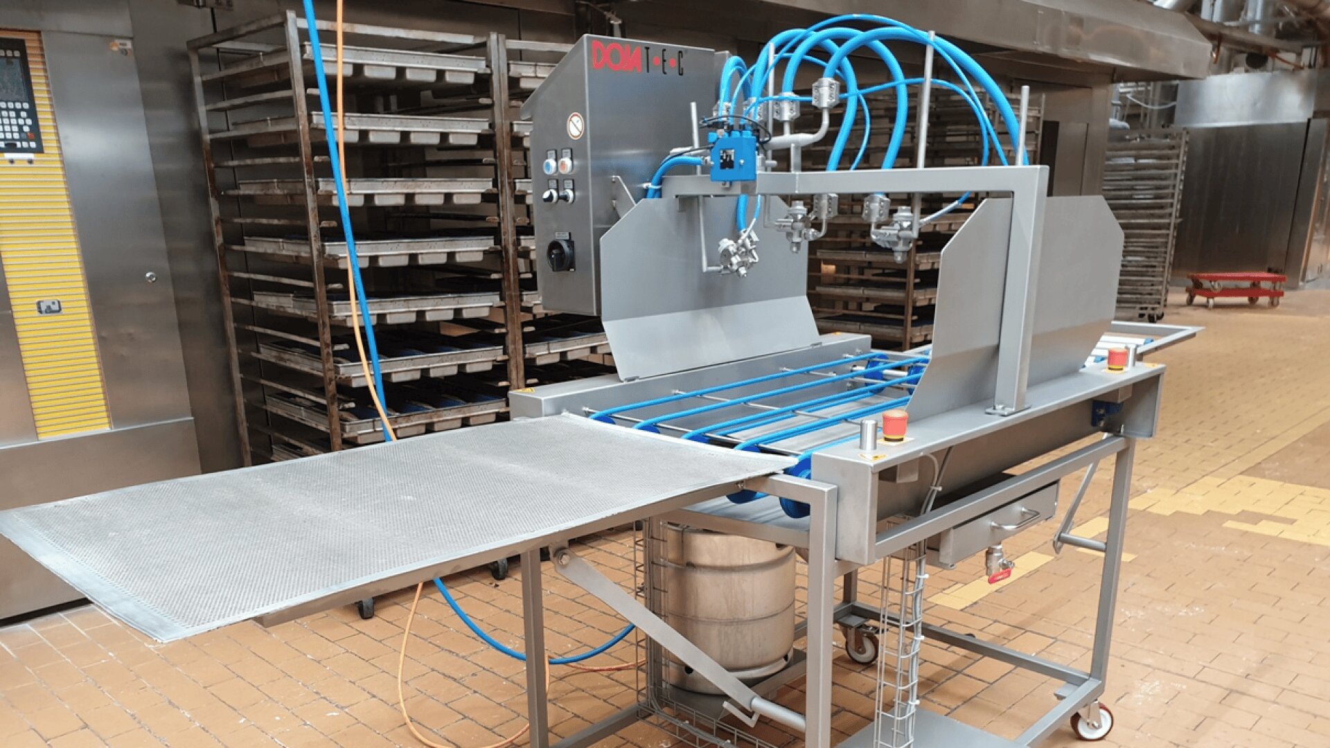 Eine industrielle Lebensmittelverarbeitungsmaschine aus Edelstahl mit blauen Rohren und Bedienfeld, die in einer Bäckerei mit Backblechen und -gestellen im Hintergrund steht.