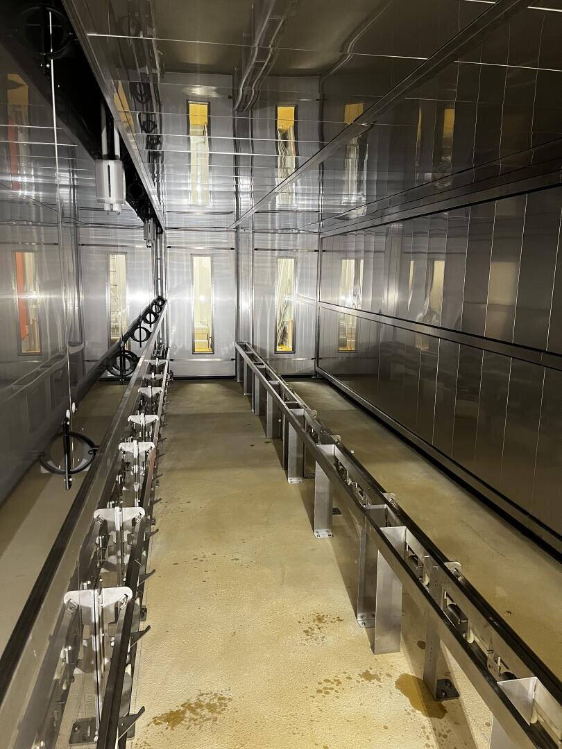 A brightly lit, narrow industrial room with shiny metallic walls and ceiling, and metal rails running along both sides of the concrete floor. There are vertical window panels at the far end that let in some light.