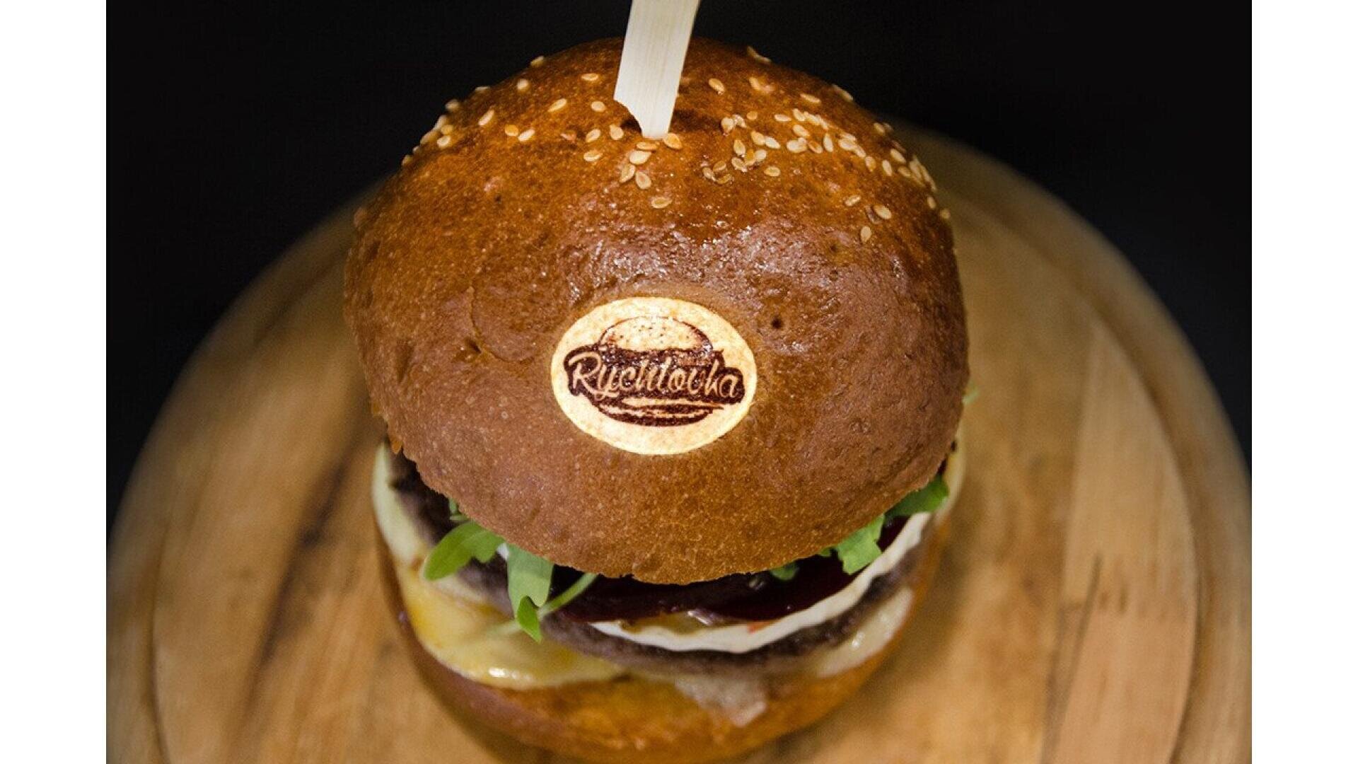 A close-up of a gourmet burger on a wooden board, topped with a sesame seed bun branded with the text Riccitella, and held together by a wooden skewer. Lettuce, cheese, and sauce are visible inside.