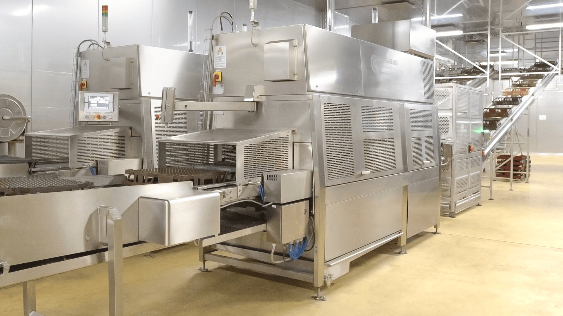 Zwei große Industriemaschinen aus rostfreiem Stahl in einer Lebensmittelverarbeitungsanlage mit Förderbändern und Schalttafeln stehen auf einem gelben Boden in der Nähe von Metalltreppen und weiteren Geräten im Hintergrund.