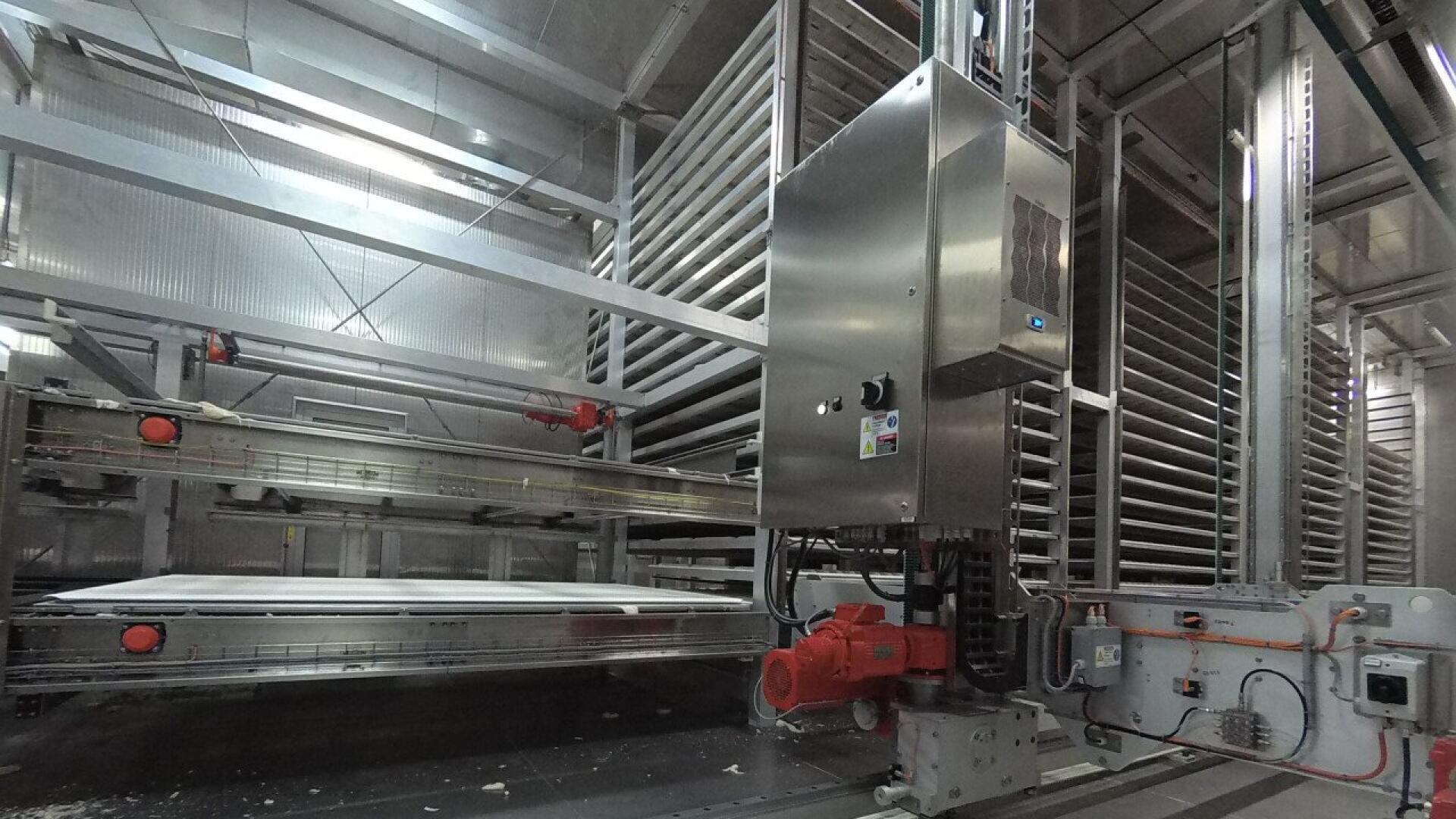Große Industriemaschine mit mehreren Metallgestellen, roten Roboterarmen und elektrischen Schalttafeln in einer modernen Fabrikumgebung. Die Umgebung ist sauber und gut beleuchtet mit reflektierenden Oberflächen.