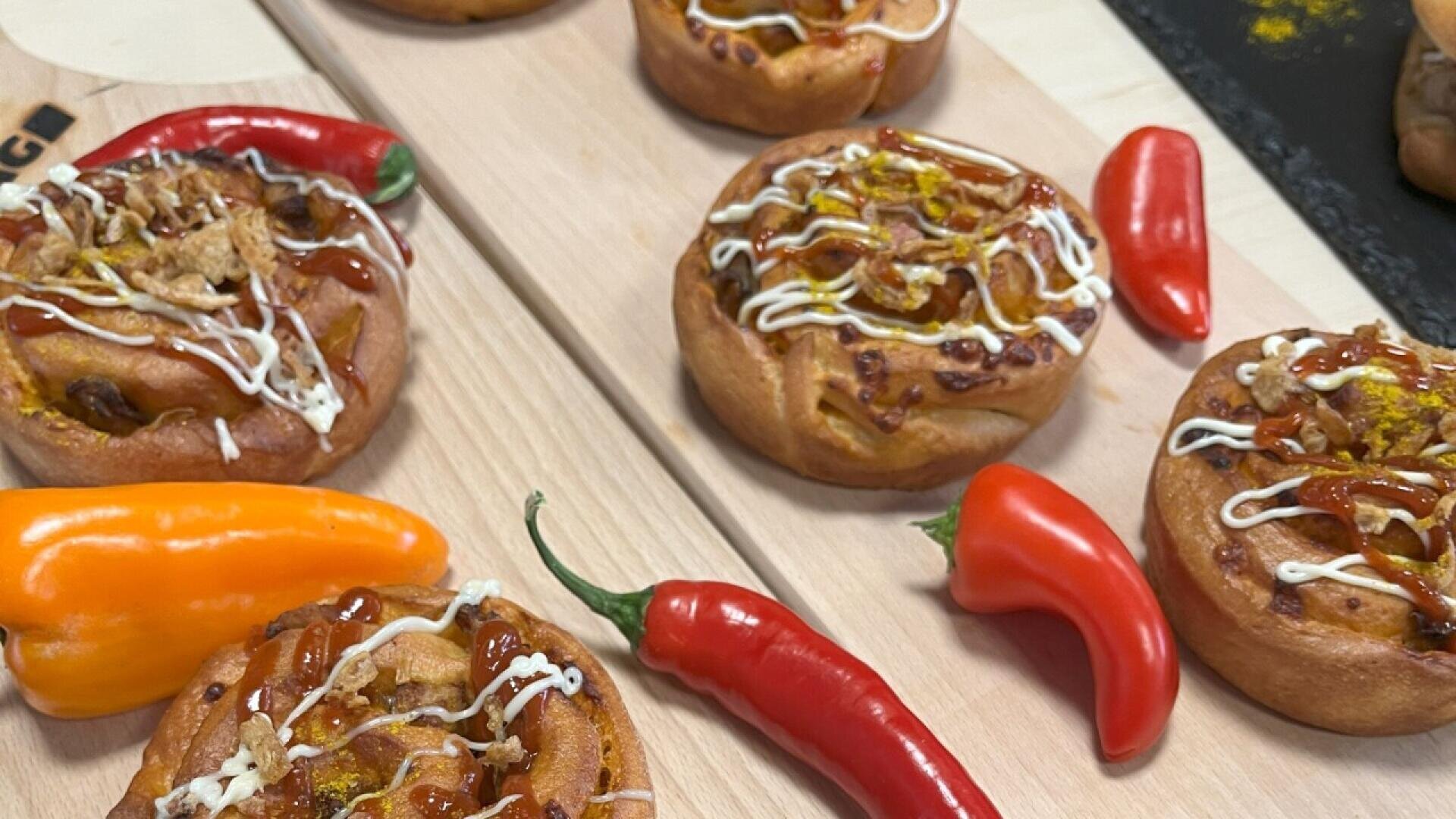 Several savory rolls topped with drizzles of sauces are displayed on wooden boards, surrounded by various colored chili peppers, including red and orange varieties.