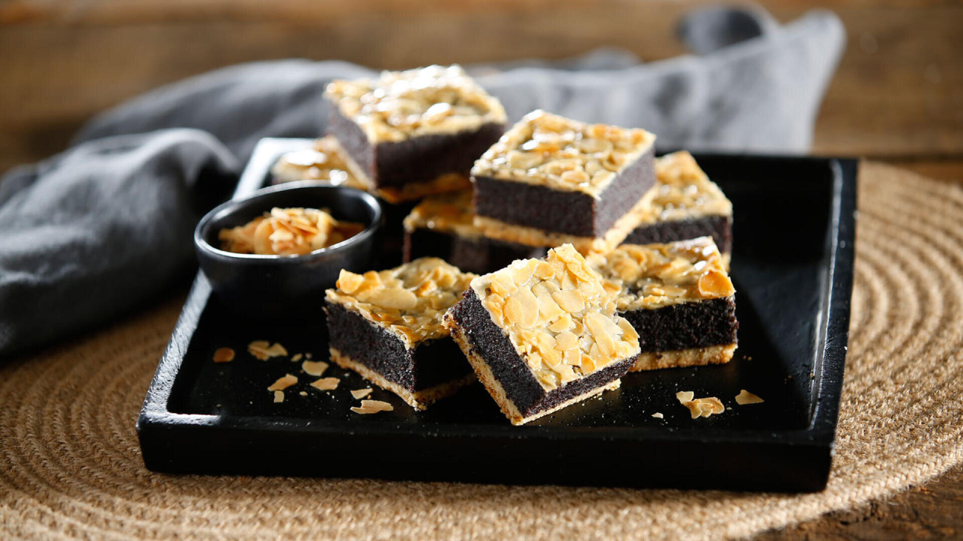 Auf einem schwarzen Tablett liegen mehrere quadratische Stücke Mohnkuchen mit gehobelten Mandeln, daneben eine kleine schwarze Schale mit Mandelblättchen, die auf einem gewebten Tischset mit einem grauen Tuch im Hintergrund steht.