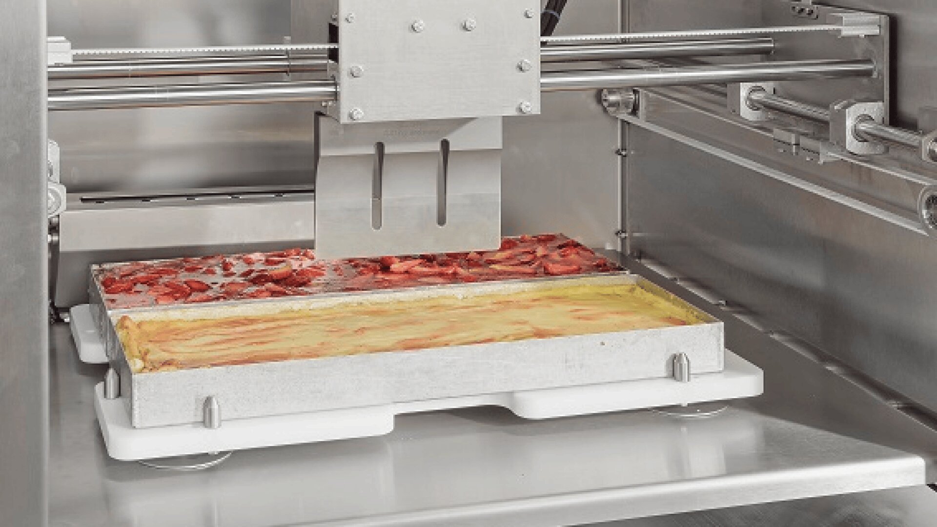 Nahaufnahme einer Lebensmittelverarbeitungsmaschine, die Schalen mit Backwaren schneidet, von denen eine mit geschnittenen Erdbeeren und die andere mit einer glatten gelben Oberfläche versehen ist. Die Schalen sind nebeneinander in der Edelstahlmaschine angeordnet.