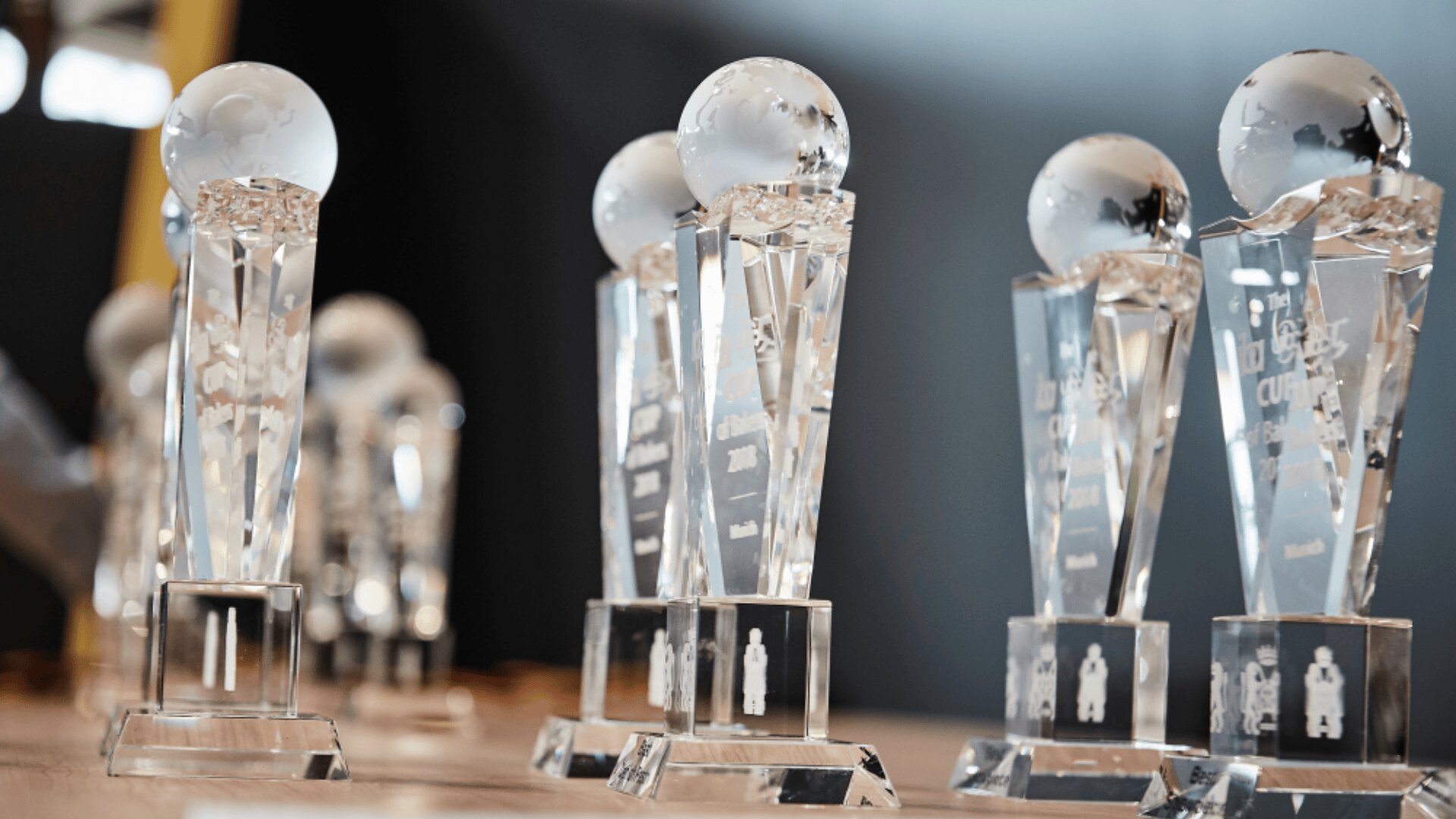 Mehrere Trophäen aus klarem Kristall mit kugelförmigen Aufsätzen sind auf einer hölzernen Oberfläche mit unscharfem Hintergrund ausgestellt. Die Trophäen haben eingravierte Texte und Motive, die das Licht reflektieren.