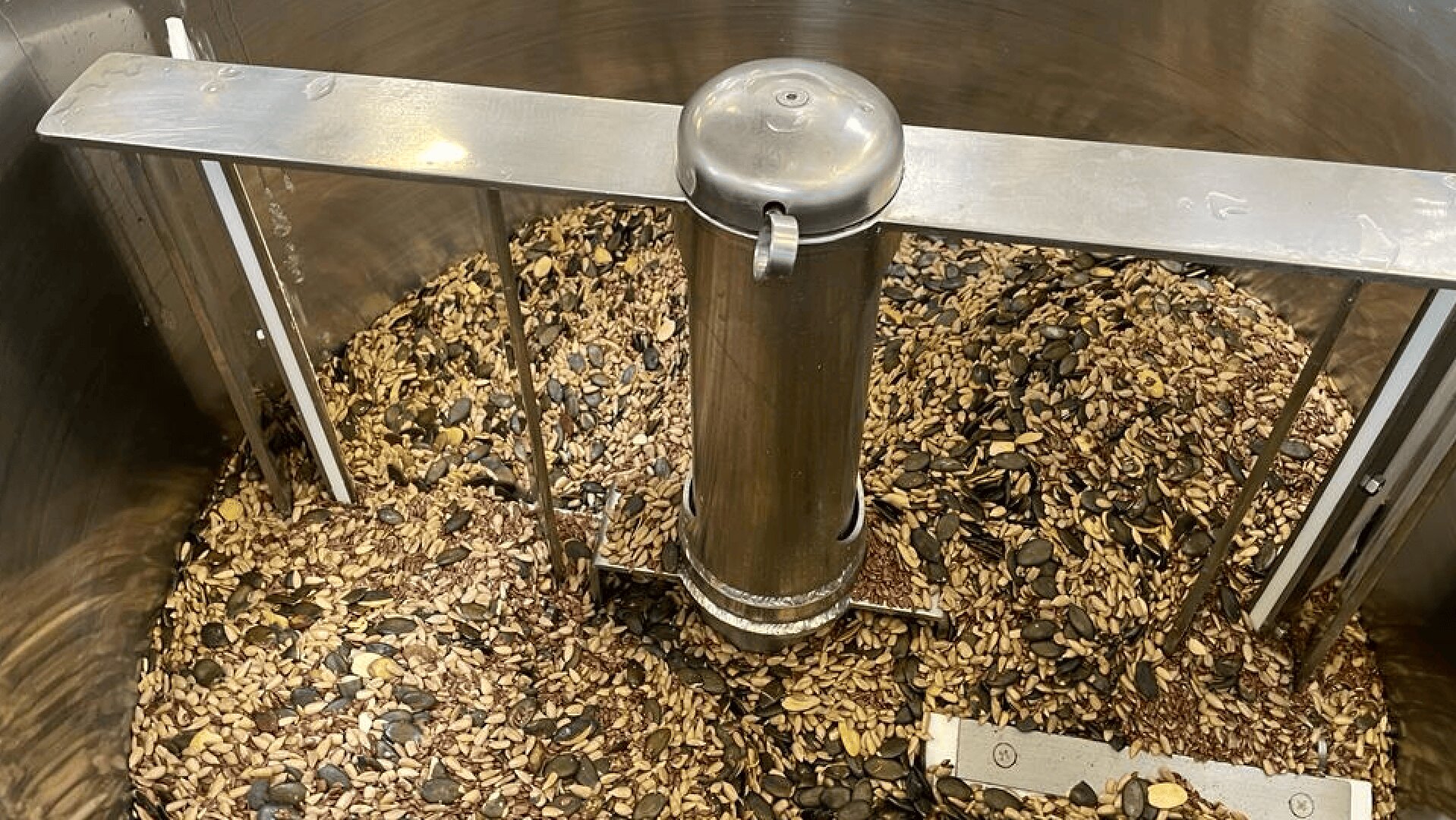 A metal mixing machine is filled with a variety of seeds, including pumpkin and sunflower seeds, being stirred together in a large stainless steel container.