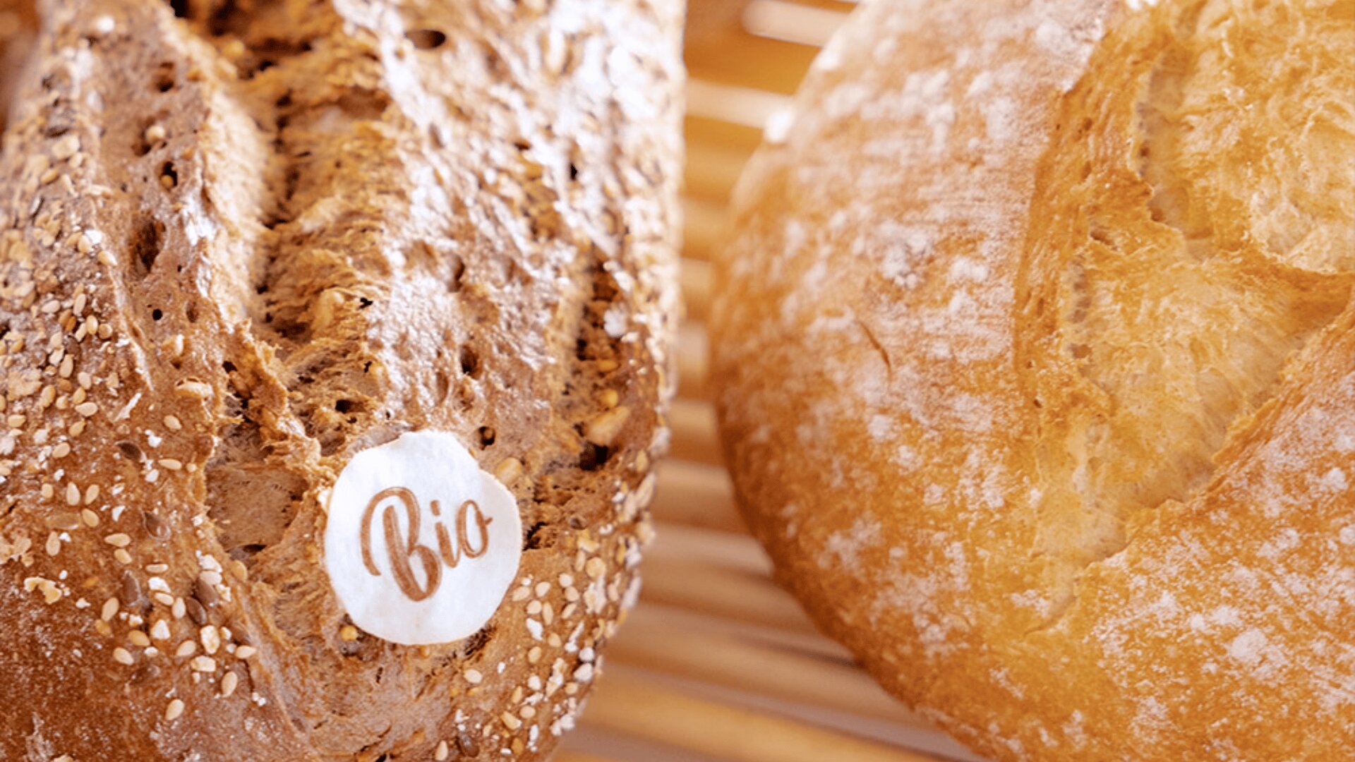 Two loaves of bread on a wooden rack; one is a seeded brown bread with a “Bio” label, and the other is a round, rustic white loaf dusted with flour.