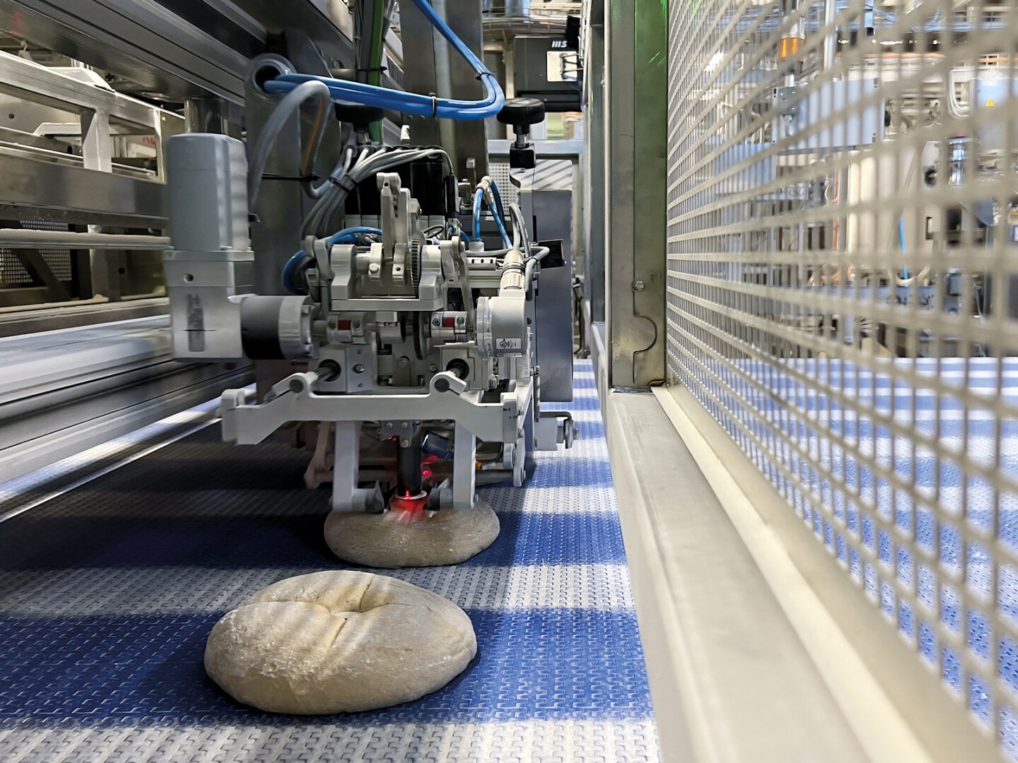 A robotic machine with mechanical parts and cables places dough pieces on a blue conveyor belt in an industrial bakery setting, with metal fencing visible on the right.