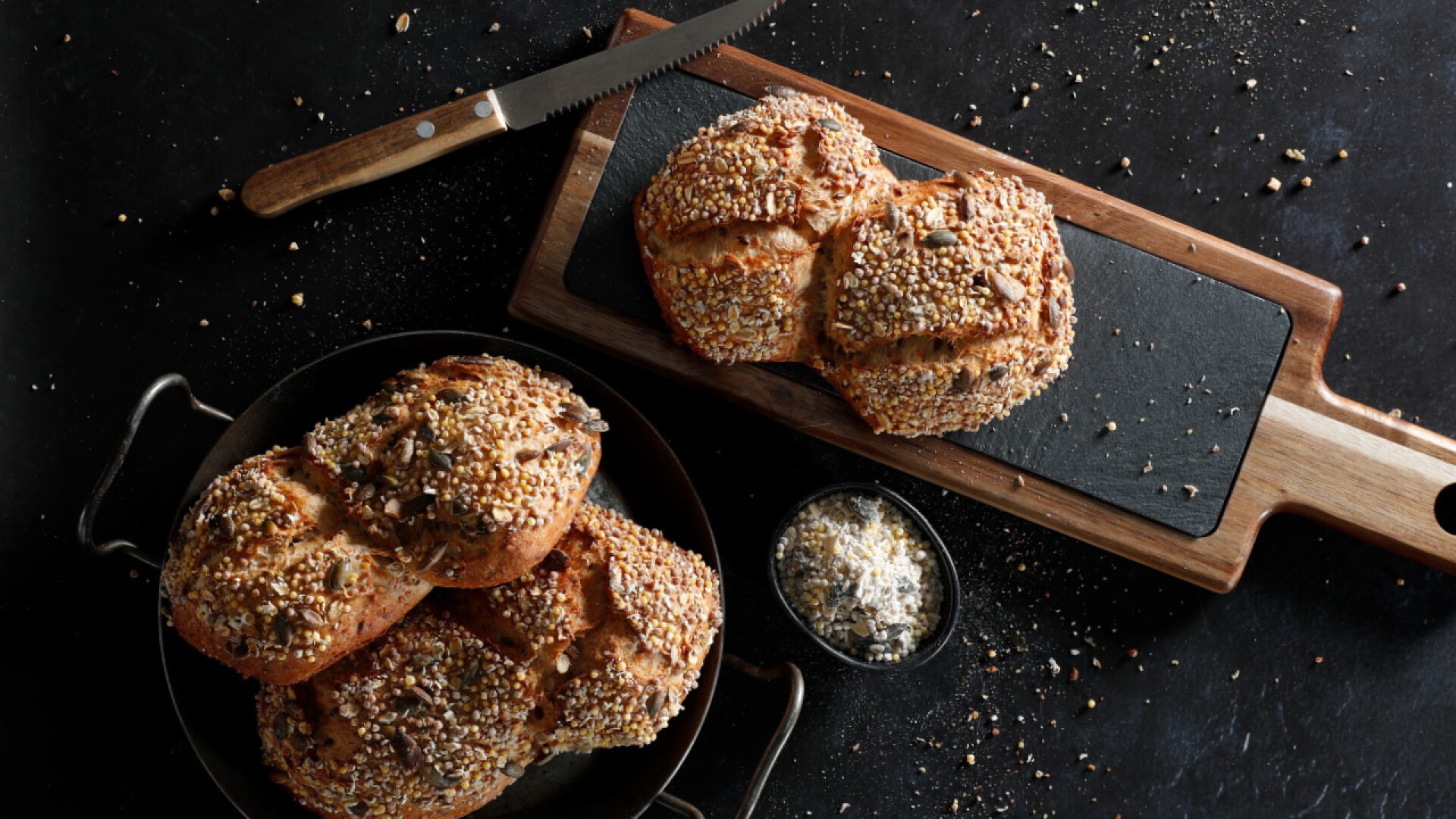 A loaf of multigrain bread on a black cutting board with a serrated knife, next to a bowl and a pan filled with more multigrain bread, all on a dark tabletop sprinkled with seeds.
