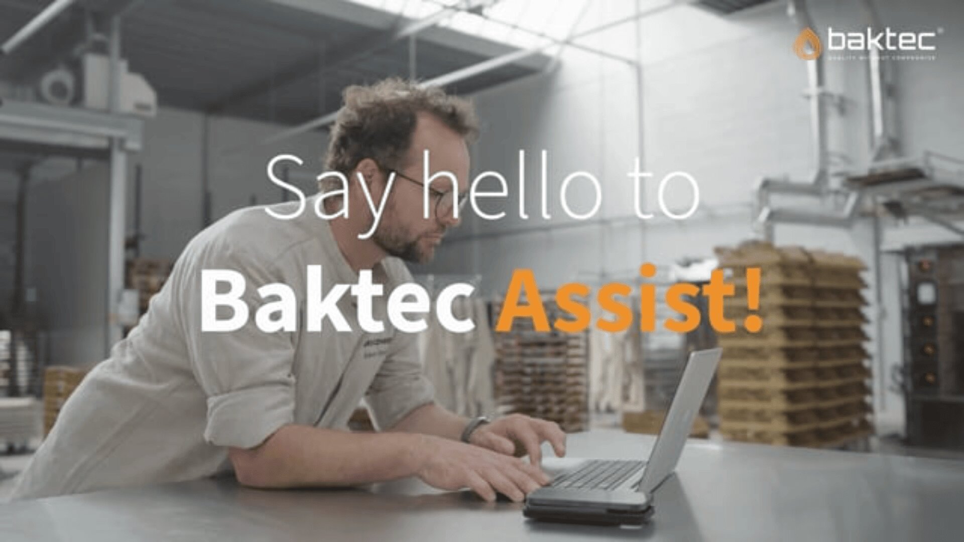 Ein Mann mit Brille und hellem Hemd arbeitet an einem Laptop in einer Bäckerei. Im Hintergrund sind Stapel von Backblechen zu sehen. Der überlagerte Text lautet: "Say hello to Baktec Assist!" mit dem Baktec-Logo oben rechts.