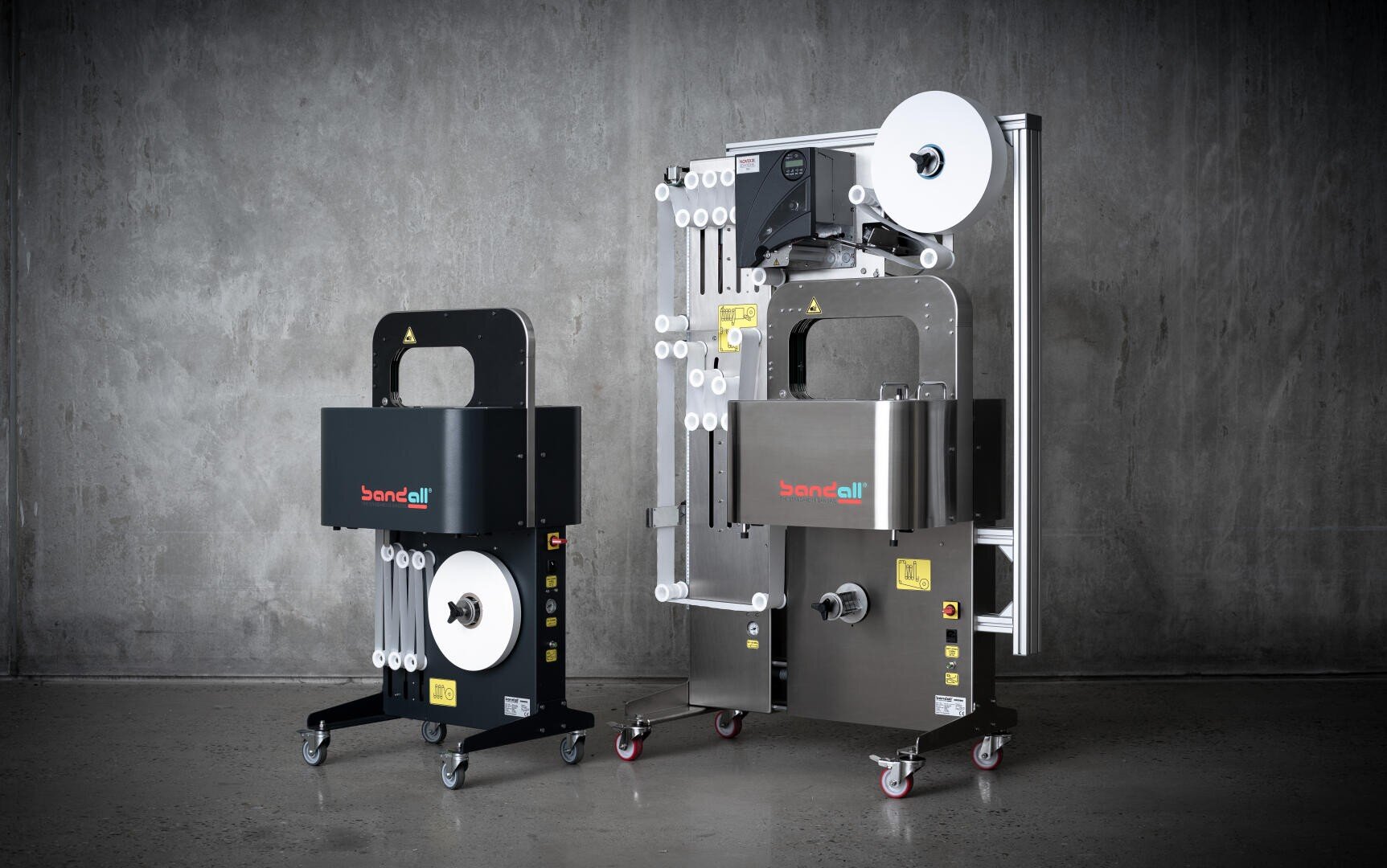 Two industrial banding machines with wheels, labeled bandall, stand side by side against a gray concrete wall. One is black and compact, the other is larger and metallic with extended rollers and a large roll of white banding material.