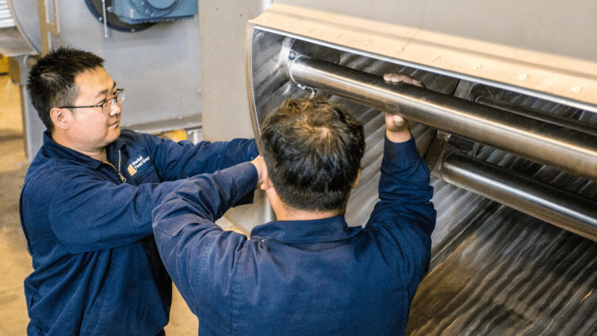 Zwei Männer in blauen Arbeitsuniformen inspizieren oder reparieren das Innere einer großen, industriellen Metallmaschine mit zylindrischen Komponenten. Sie wirken konzentriert und arbeiten gemeinsam in einer industriellen Umgebung.
