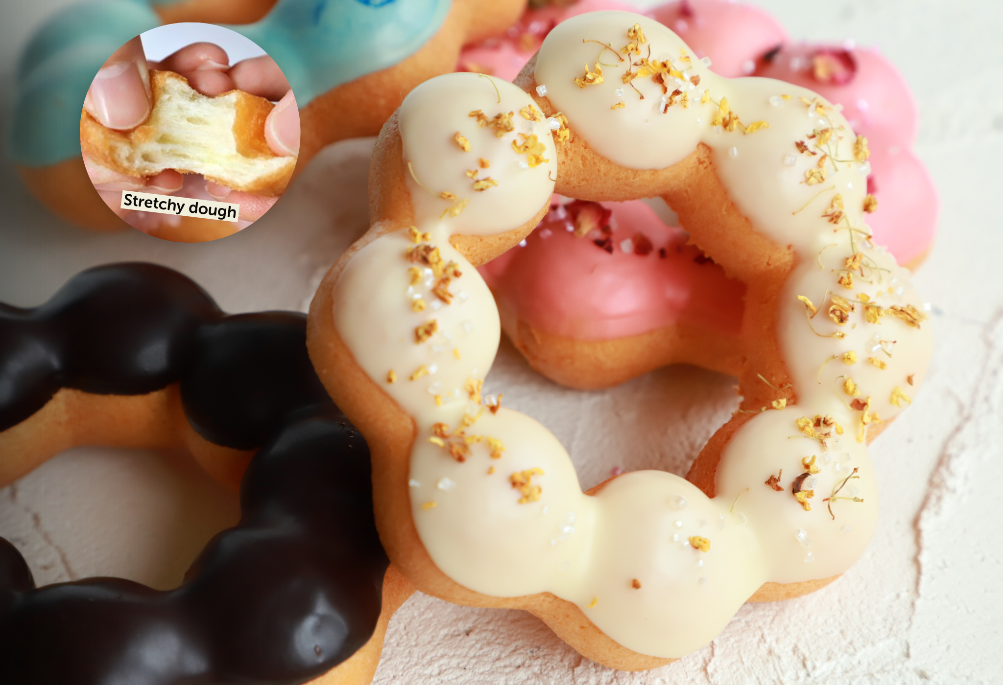 Eine Nahaufnahme von bunten, glasierten Mochi-Donuts, die auf einer hellen Fläche angeordnet sind. Eine Einblendung zeigt eine Hand, die einen Donut auseinanderzieht und den dehnbaren Teig mit der Aufschrift Stretchy dough hervorhebt.