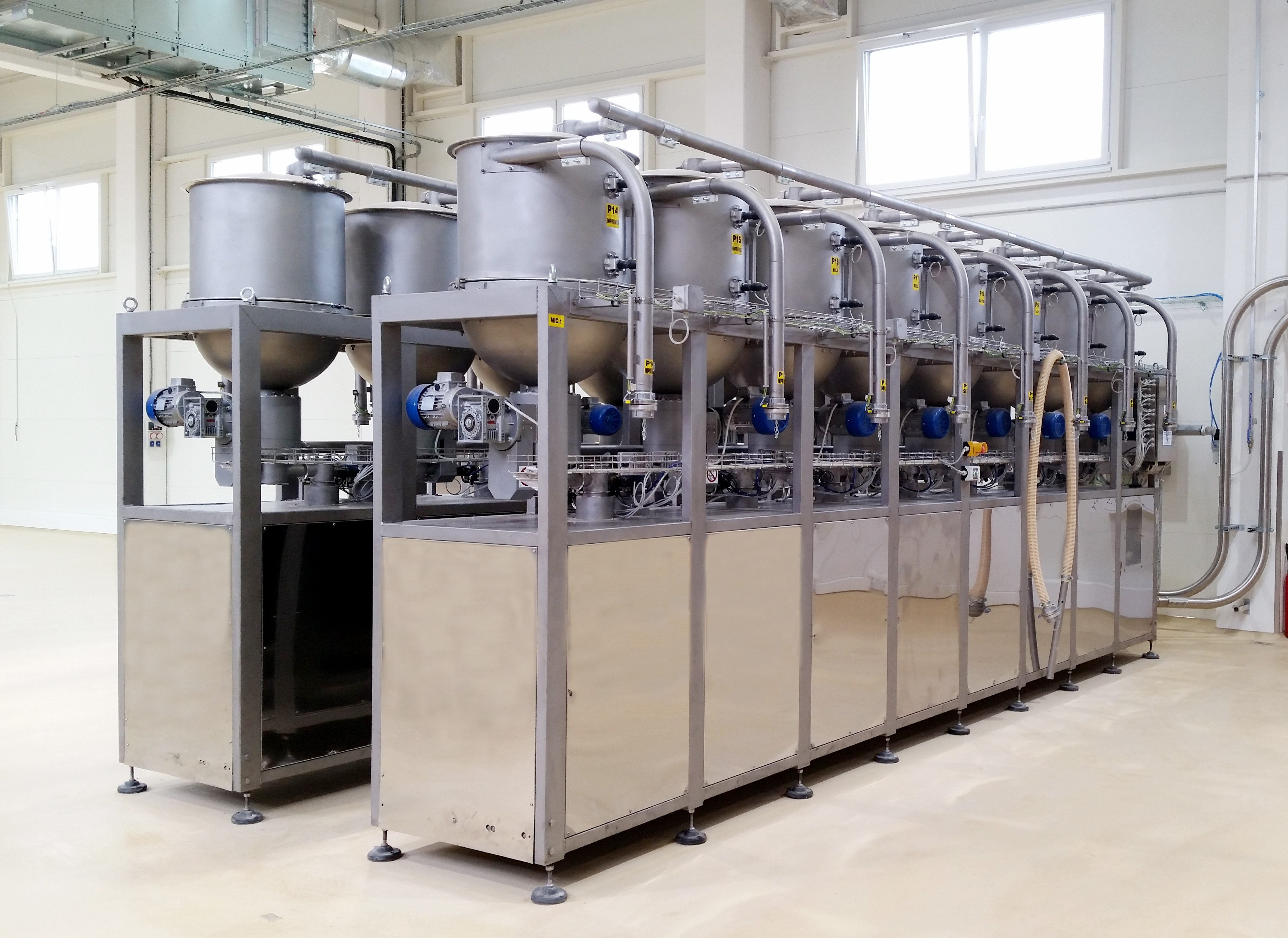 A large, industrial stainless steel machine with multiple tanks, pipes, and motors stands in a clean, bright factory room with cream-colored floors and white walls.