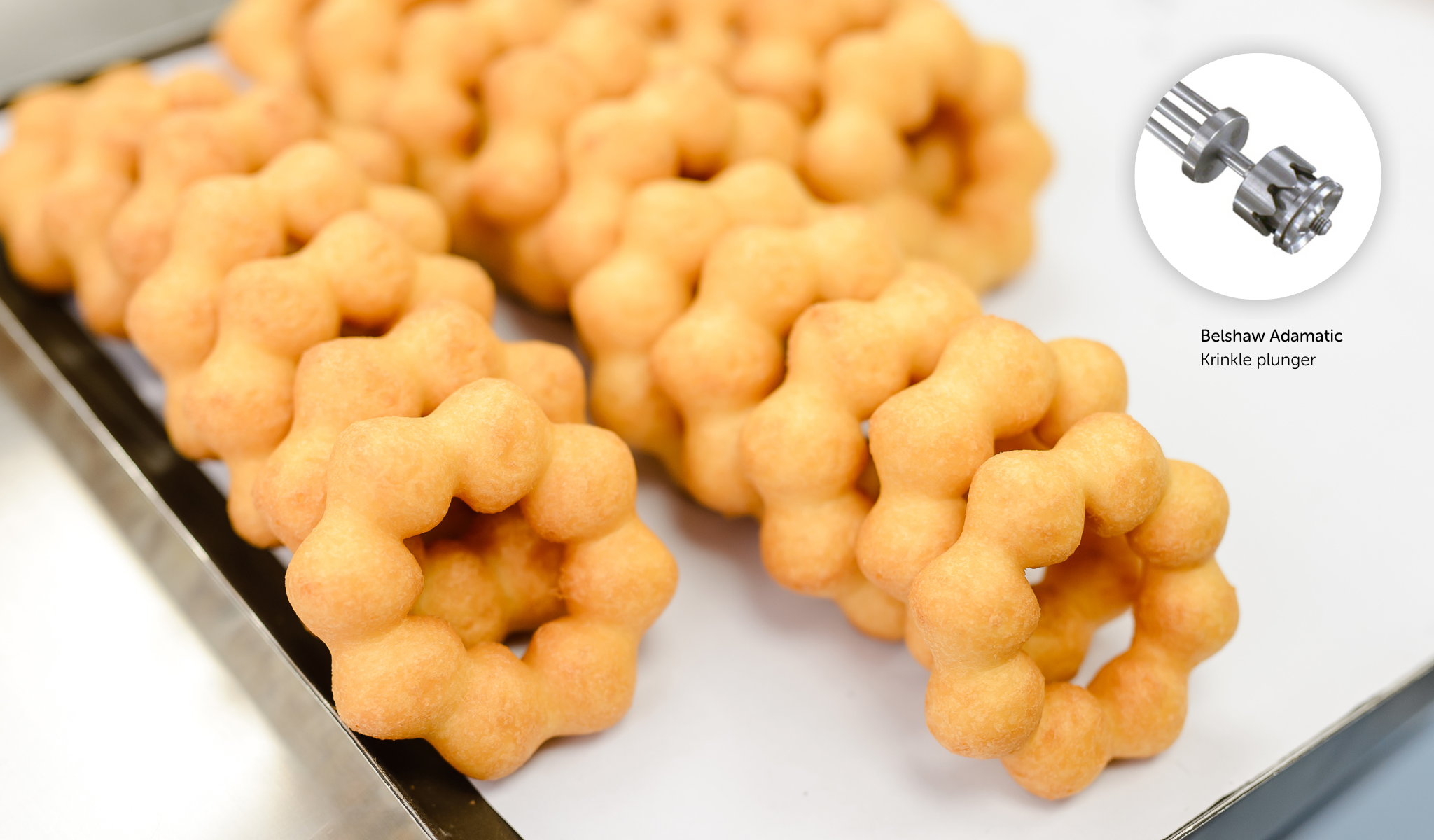 Reihen von goldenen, ringförmigen Donuts mit blasenartigen Segmenten auf einem Tablett. Inset zeigt eine Nahaufnahme eines Metall-Donutschneiders mit der Bezeichnung Belshaw Adamatic Krinkle Plunger.