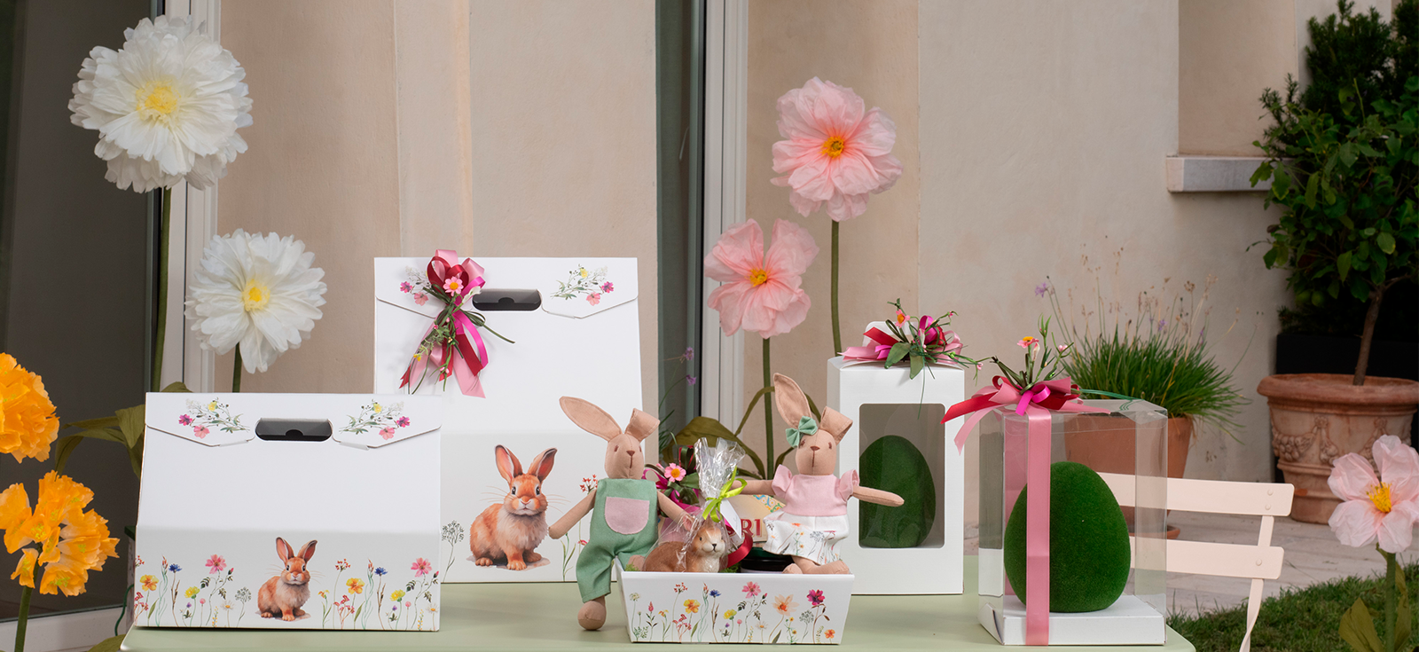 Eine festliche Frühlingsdekoration mit Geschenkboxen mit Hasenmotiven, Plüschhasen, rosa und weißen Papierblumen, einem grünen, eiförmigen Ornament und einem kleinen Holzkarren, alles arrangiert auf einem hellgrünen Tisch im Freien.