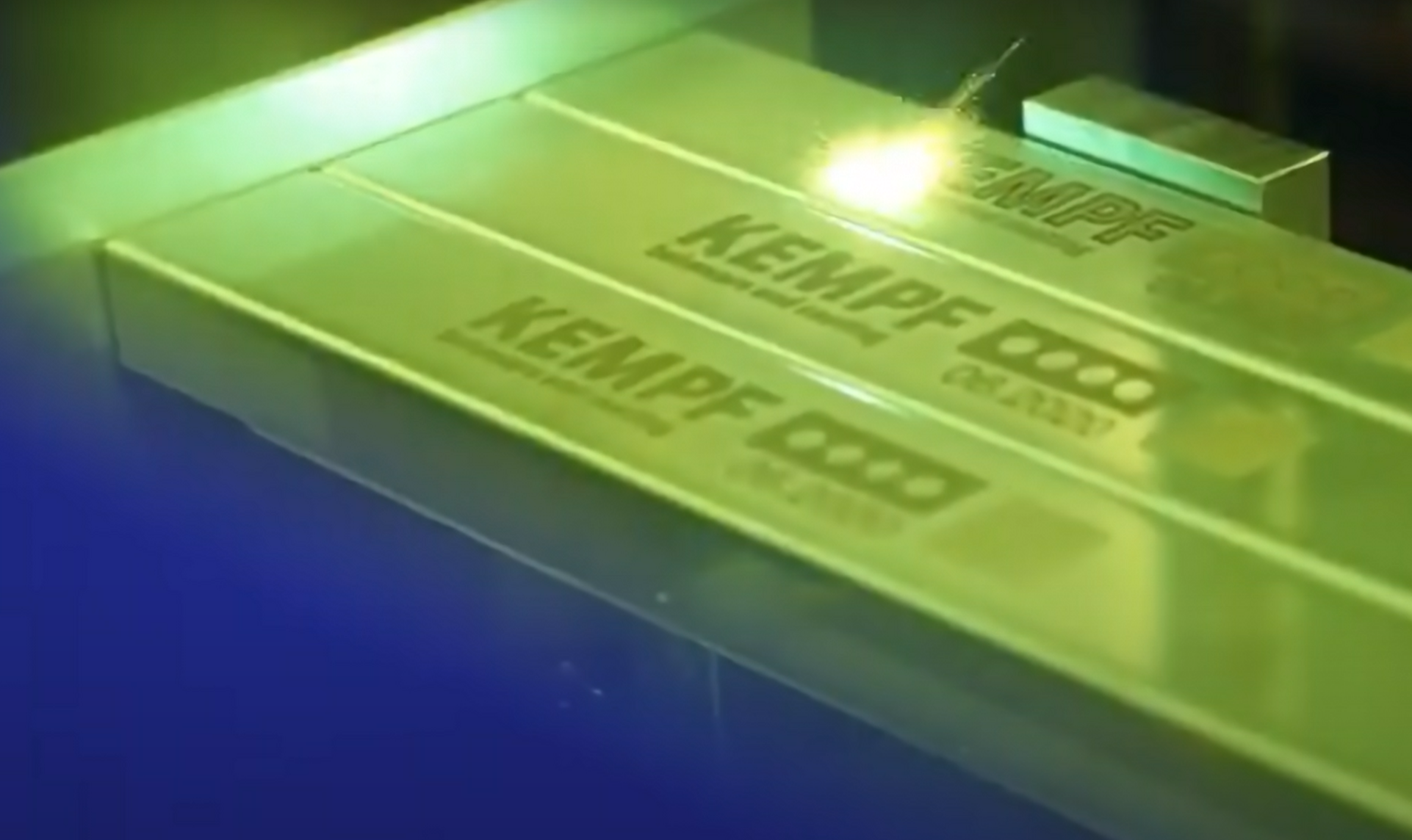 Ein Laser ätzt den Text "KEMPF" und Symbole in einen Metallbarren. Zwei fertige Stäbe mit demselben Design sind neben der laufenden Gravur zu sehen.