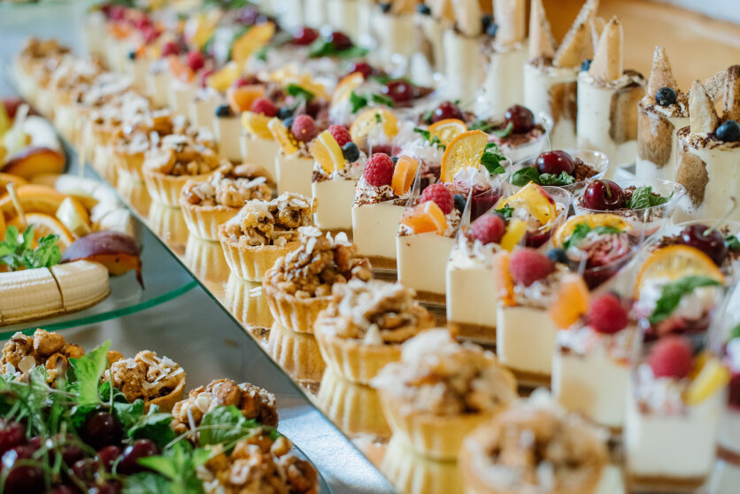 Eine farbenfrohe Auslage mit verschiedenen Mini-Desserts, darunter Törtchen mit Nüssen, mit Sahne gefüllte Becher mit Früchten und dekorative Kuchen, die ordentlich auf einem Büffettisch angeordnet sind.