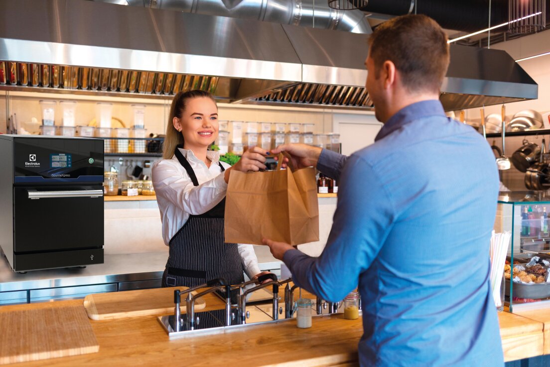 Eine lächelnde Frau in weißem Hemd und Schürze reicht einem Mann über die Theke einer modernen Küche oder eines Cafés hinweg eine Papiertüte. Im Hintergrund sind Edelstahlgeräte und Regale mit Gläsern zu sehen.