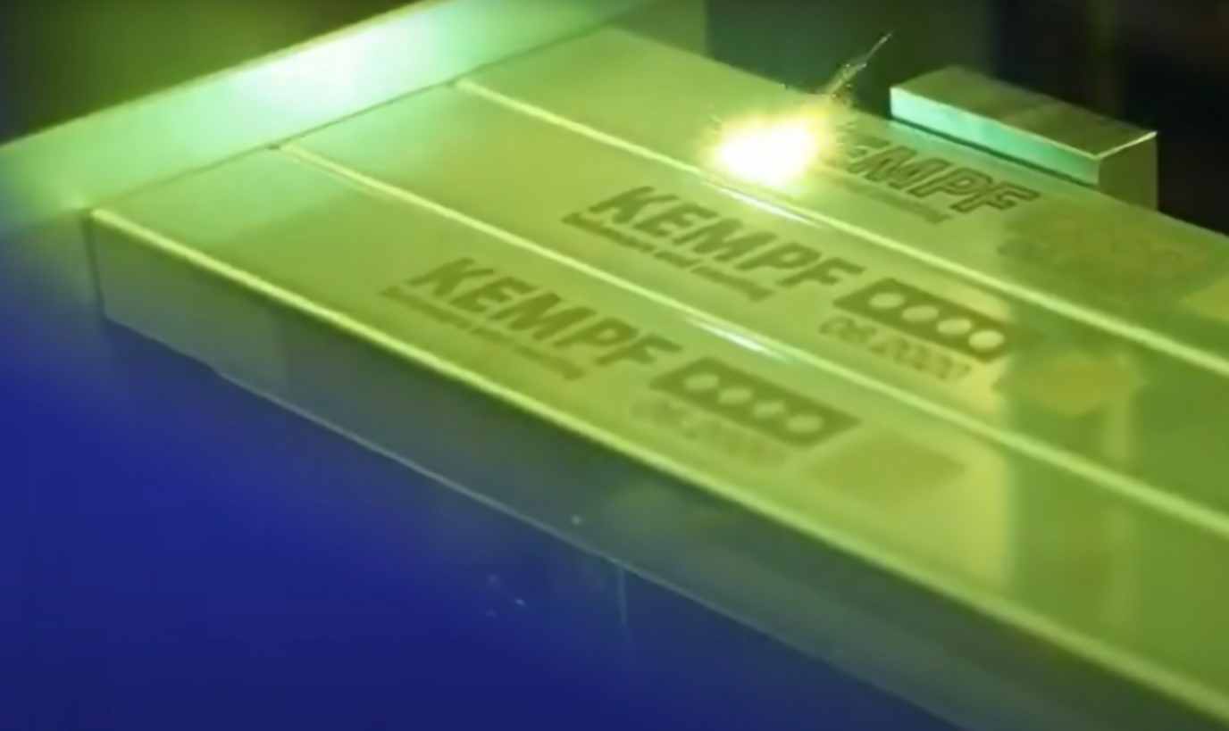 A laser engraves the word KEMPF and symbols onto a metal surface, with a bright yellow light shining at the point of contact.