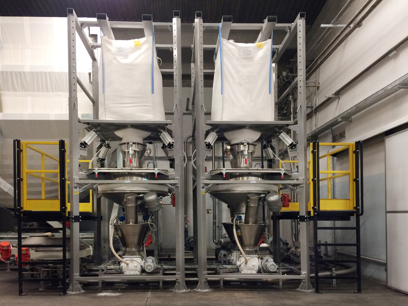 Two large industrial machines with metal frames and yellow safety railings hold big white bulk bags at the top, connected to funnel-shaped hoppers below, inside a clean, modern factory setting.